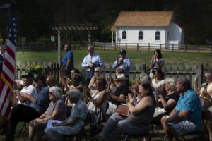 Outdoor audience clapping at community gathering near small white chapel under sunny skies with American flag visible.