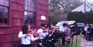 String quartet performs outdoors beside red building with audience seated in background.