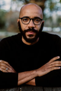 Clint Smith with glasses and beard sitting outdoors, arms crossed, wearing a black shirt.