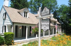 Historic Wayside Cottage with garden, colonial architecture, sign, and American flag under clear blue sky.