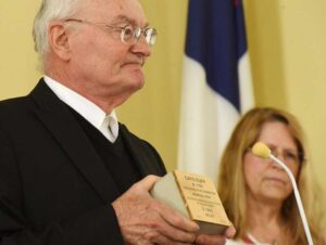 Man holding an award at a ceremony with a woman and flag in the background.