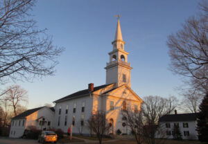 Historic white church with a tall steeple under a blue sky during sunset, surrounded by bare trees.