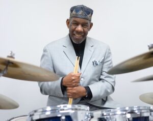 Elderly drummer in a suit smiles while holding drumsticks, sitting behind a drum kit, wearing a patterned hat.