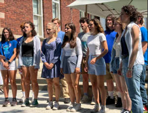 A group of young people singing outdoors in casual summer attire in front of a brick building.
