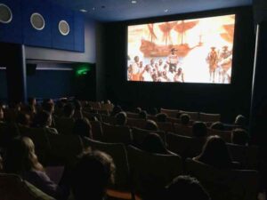 Audience watching a historical film in a dimly lit theater, with a bright screen displaying a ship scene.