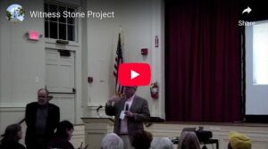 Speaker presenting at the Witness Stone Project event in a hall with an audience, flag, and projection screen in view.