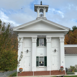 White historical building with green shutters and bell tower, set against autumn trees and a cloudy sky.