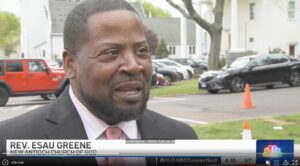 Man in a suit speaking outside, parked cars and orange cone visible in the background.