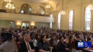 Crowded church hall during an event, filled with attentive attendees seated and standing, with chandeliers above.