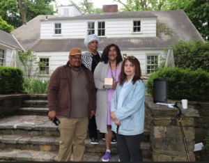 Group of people posing outside a white house with plaque, surrounded by greenery and stone steps.