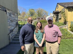 Three people smiling outdoors in a garden setting near rustic buildings under a clear blue sky.