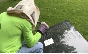 Worker engraves Revolutionary War gravestone on grass-covered ground.