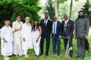 Group photo of people dressed in formal and casual attire, outdoors with lush greenery and a saxophonist.