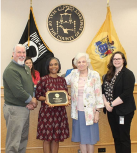 Award ceremony in Somerset County with a diverse group posing around a plaque. Flags and seal visible in background.