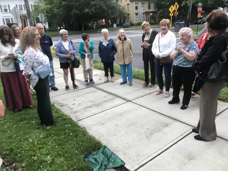 A group of people gathers on a sidewalk, engaging in conversation near a residential street.