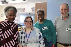 A group of four adults smiling together in a classroom setting, promoting community and education.