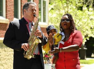 Man playing saxophone, woman singing with child outside on sunny day, capturing joyful musical moment.