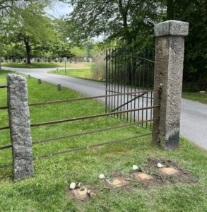 Rustic gate opening to a serene cemetery path surrounded by greenery and gravestones in the background.