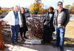Group of people smiling outdoors on a sunny day, standing near a plant with autumn foliage in the background.