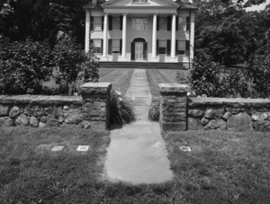 Historic colonial-style house with columns and stone path, surrounded by lush gardens and stone walls.