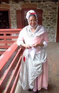 Person in historical attire, bonnet and glasses, stands on a wooden porch with stone building in background.