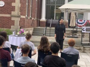 Speaker addressing an audience outdoors at an educational event with patriotic decorations and historic building backdrop.