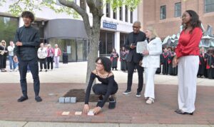 Person placing commemorative brick on sidewalk during group event with attendees and choir in the background.