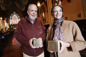 Two people smiling and holding engraved plaques inside a beautiful, historic church with stained glass windows.