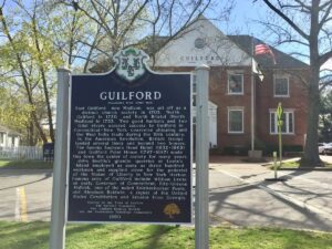 Historic Guilford sign with detailed history text, located in front of a red brick building and trees.
