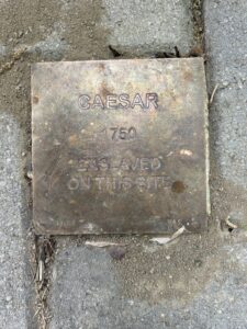 Bronze plaque on pavement marking historical site where Caesar was enslaved in 1750.