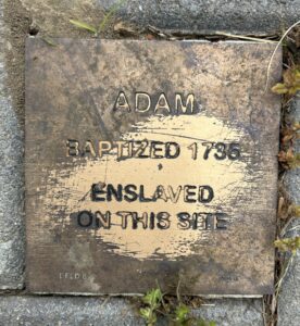 Bronze plaque reading Adam, Baptized 1735, Enslaved on this Site with weathered texture and surrounding grass.