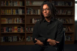 Person sitting in a library, wearing black, with bookshelves in the background.