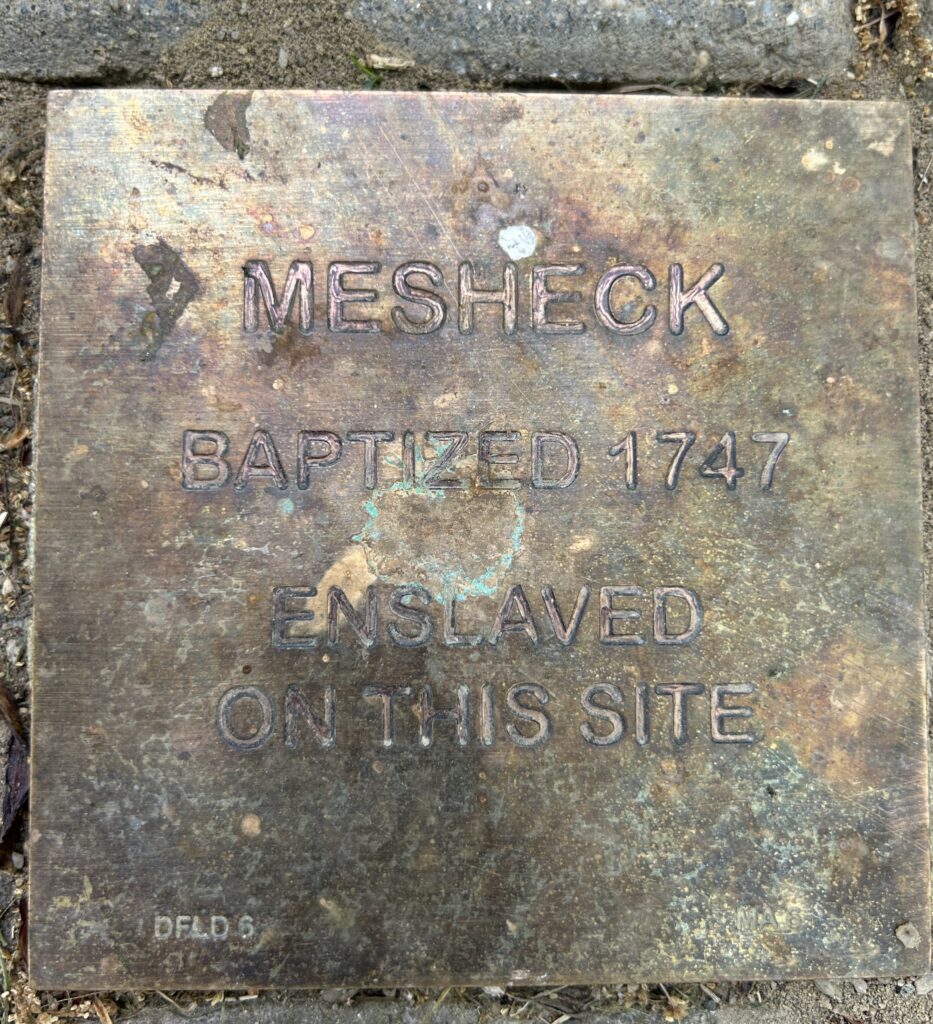 Historic plaque for Mesheck, baptized 1747, marking site of enslavement. A reminder of past injustices.