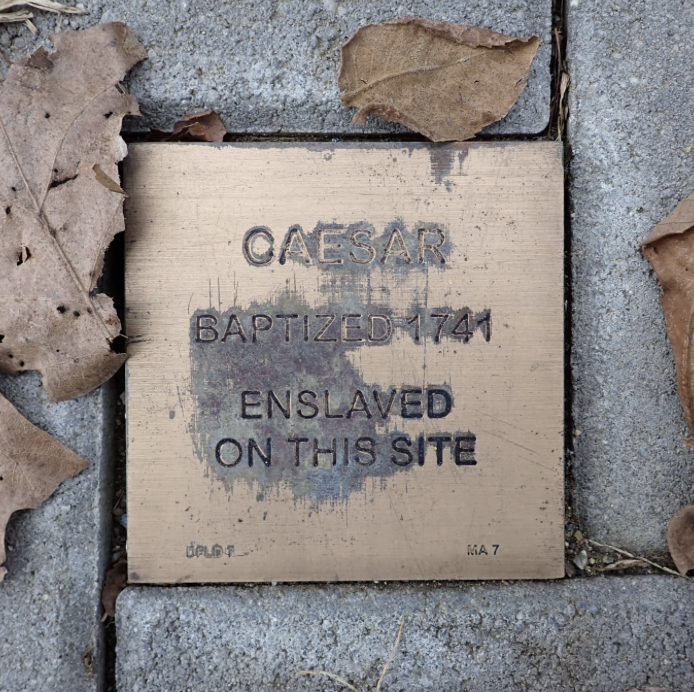 Memorial plaque for Caesar, baptized 1741, enslaved, amid fallen leaves on cobblestone.