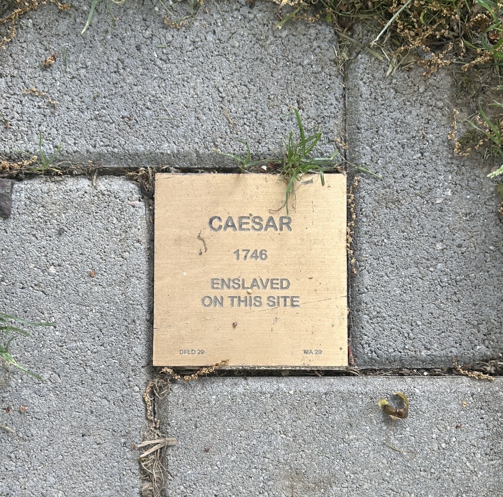 Commemorative plaque on pavement for Caesar, enslaved in 1746, highlighting historical site.