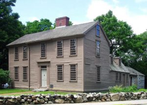 A photo of the exterior of the Hancock Clarke House in Lexington, Massachesetts. The leaves on the trees are green and it is a partially cloudy day with blue skies.