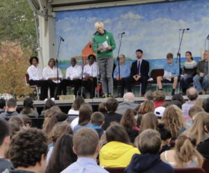A student in a green sweatshirt, with blond hair, giving a speech on a stage in front of a crowd.