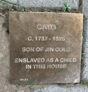 Bronze plaque honoring Cato, son of Jin Cole, enslaved as a child, 1737-1825, found on the ground with grass.