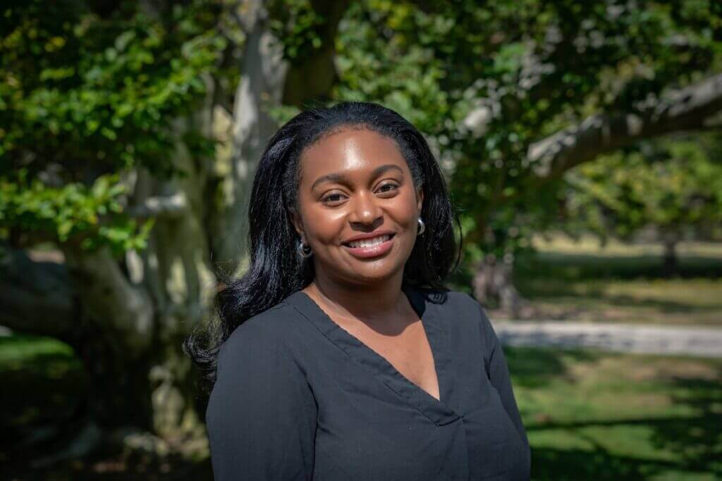 Smiling woman in a black top posing outdoors with green trees in the background on a sunny day.