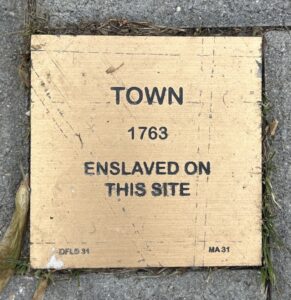 Historical plaque on pavement reading Town 1763 Enslaved On This Site in bold black text.