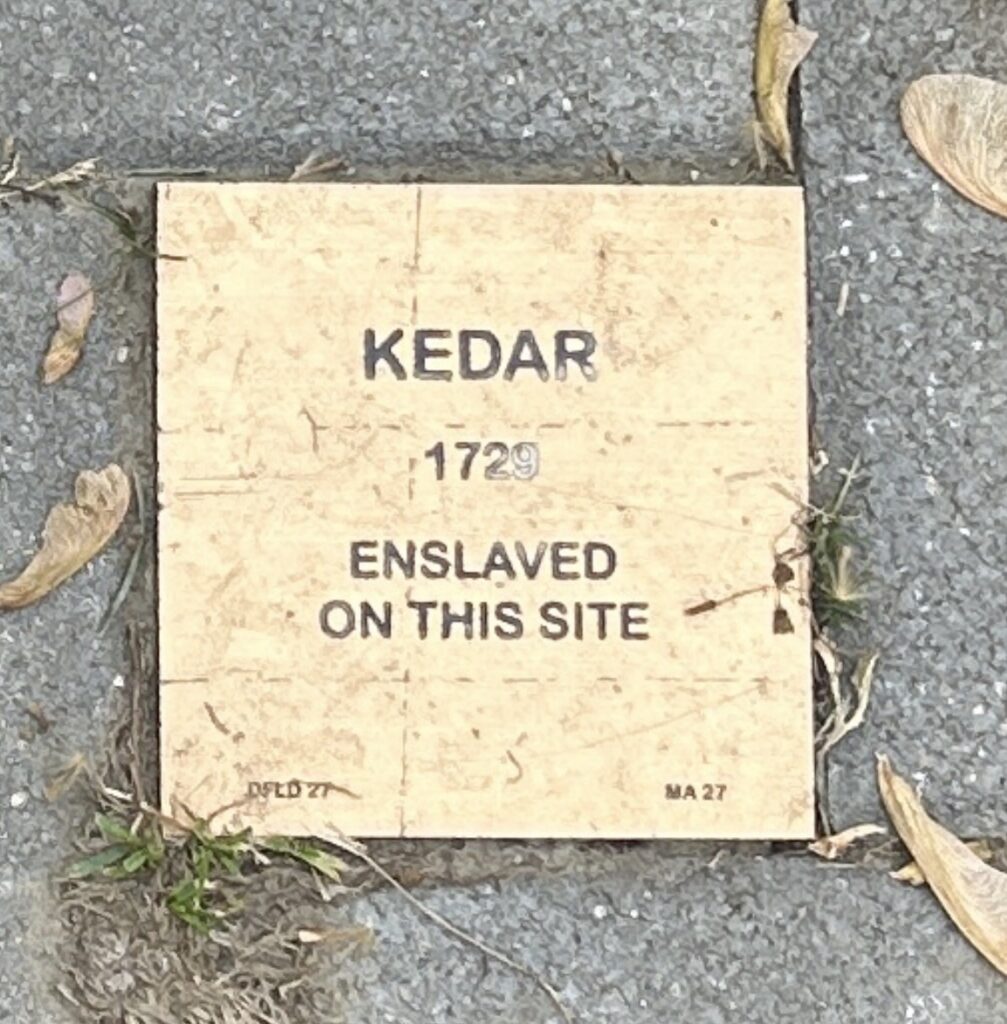 Memorial plaque for Kedar, enslaved in 1729, set in a sidewalk surrounded by leaves and grass.
