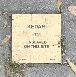 Memorial plaque for Kedar, enslaved in 1729, set in a sidewalk surrounded by leaves and grass.