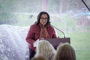 Woman speaking outdoors at a podium, engaging audience in a red jacket and scarf, with nature background.