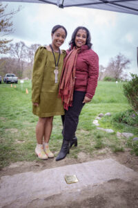 Two women smiling at a garden ceremony, standing beside a commemorative plaque on the ground.
