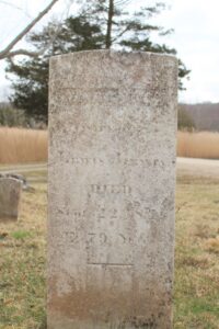 Old weathered gravestone in a cemetery, surrounded by grass and trees in the background.
