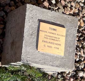 Memorial plaque for Tome, a mason, farmer, builder, enslaved from 1682-1708, on a concrete block amidst stones.