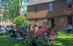 Outdoor lecture at historic brown house with audience in folding chairs, surrounded by green trees and sunny skies.