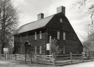 Historic colonial house with weathered wood siding, surrounded by a wooden fence and leafless trees on a cloudy day.