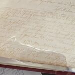 Old handwritten document in a protective sleeve, showing cursive text with dates and signatures, on a historical topic.