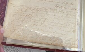 Old handwritten document in a protective sleeve, showing cursive text with dates and signatures, on a historical topic.
